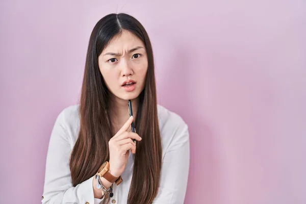 中国の若い女性はショック顔でピンクの背景に鉛筆を保持 懐疑的で皮肉を探して オープン口で驚いて — ストック写真