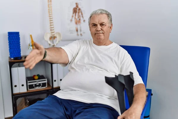 Homem Caucasiano Sênior Clínica Fisioterapia Segurando Muletas Sorrindo Amigável Oferecendo — Fotografia de Stock