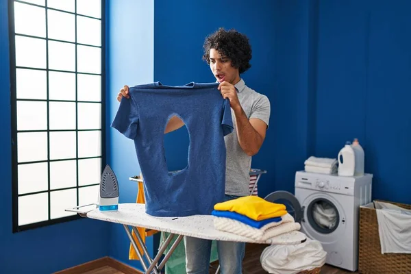 洗濯室で焼かれた鉄のシャツを保持している巻き毛のアイロンを持つヒスパニック系の男は恐れて 驚きと驚きの表情でショックを受けました 恐怖と興奮した顔 — ストック写真