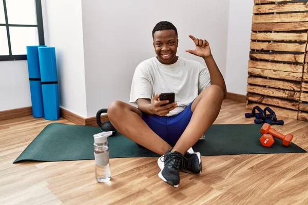 Young african man sitting on training mat at the gym using smartphone smiling and confident gesturing with hand doing small size sign with fingers looking and the camera. measure concept.