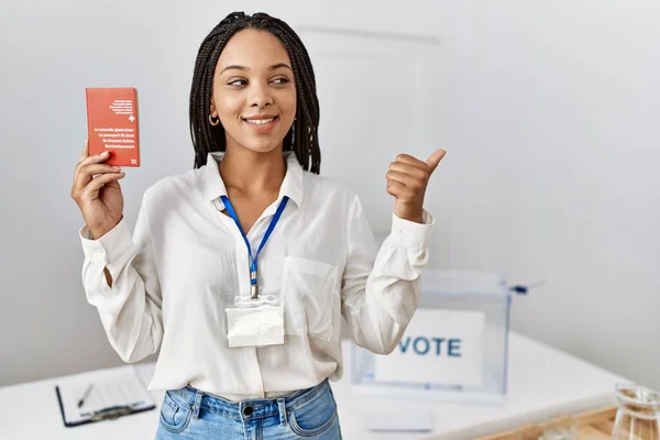 Young african american woman at political campaign election holding swiss passport pointing thumb up to the side smiling happy with open mouth