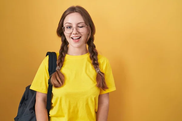 Young caucasian woman wearing student backpack over yellow background winking looking at the camera with sexy expression, cheerful and happy face.