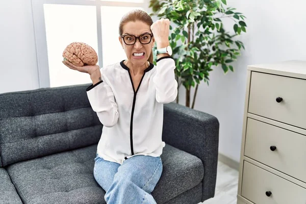 Young hispanic woman holding brain working at psychology clinic annoyed and frustrated shouting with anger, yelling crazy with anger and hand raised