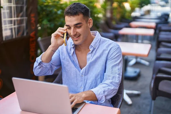 Young hispanic man using laptop talking on smartphone sitting on table at coffee shop terrace