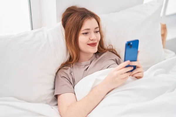 Genç Kızıl Saçlı Kadın Yatakta Akıllı Telefon Kullanıyor — Stok fotoğraf
