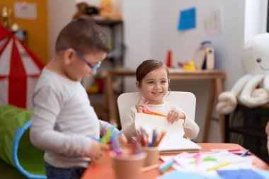 Adorable girl and boy sitting on table cutting paper at kindergarten