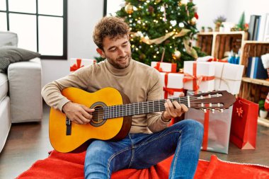 Gitar çalan genç İspanyol adam Noel ağacının yanında oturuyor.