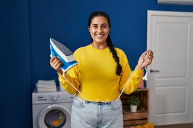 Young hispanic woman holding iron at laundry room smiling with a happy and cool smile on face. showing teeth. 