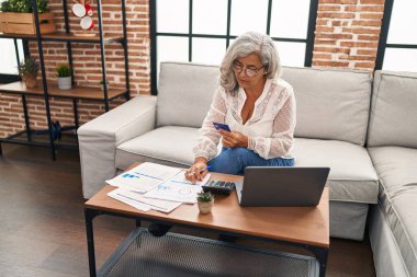Evde dizüstü bilgisayar ve kredi kartı kullanan orta yaşlı kadın.