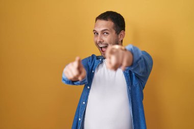 Sarı arka planda duran İspanyol adam sana ve kameraya parmaklarıyla bakıyor, gülümsüyor ve neşeli. 