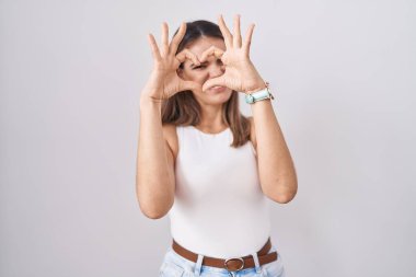 Hispanic young woman standing over white background doing heart shape with hand and fingers smiling looking through sign 