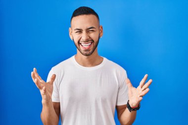 Mavi arka planda duran genç İspanyol adam başarı için deli ve çılgınlığı kutluyor kollarını kaldırmış ve gözlerini kapatmış heyecanla bağırıyor. Kazanan konsept 