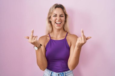 Young blonde woman standing over pink background showing middle finger doing fuck you bad expression, provocation and rude attitude. screaming excited 