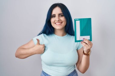 Young modern girl with blue hair holding l sign for new driver smiling happy and positive, thumb up doing excellent and approval sign 