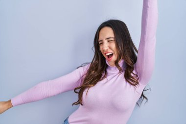 Genç esmer kadın mavi arka planda dans ediyor mutlu ve neşeli, hareketli ve kendinden emin müzik dinliyor. 