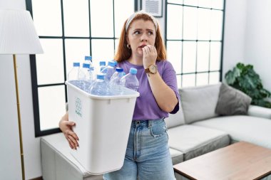 Geri dönüşüm sepetini elinde plastik şişelerle tutan genç kızıl saçlı kadın gergin ve elleri ağız yiyen tırnaklarla gergin görünüyor. anksiyete. 
