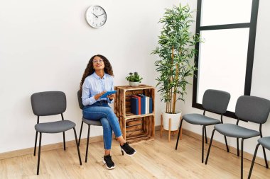 Bekleme odasında sandalyede oturan dokunmatik ped kullanan genç Latin kadın.