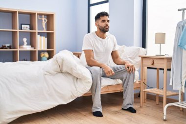 Young hispanic man with serious expression sitting on bed at bedroom