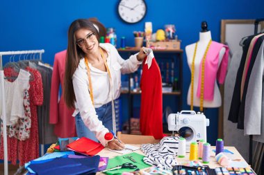 Young beautiful hispanic woman tailor holding t shirt writing on notebook at clothing factory