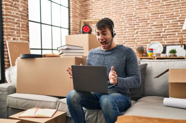 Young hispanic man working with laptop at new home celebrating victory with happy smile and winner expression with raised hands 