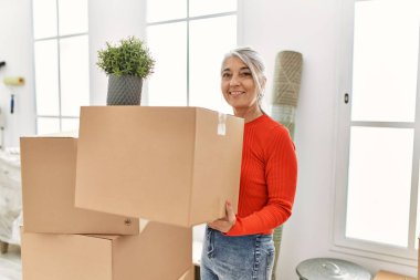 Orta yaşlı, kır saçlı, gülümseyen bir kadın yeni evinde kendine güvenen bir paket tutuyor.