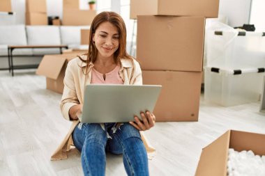 Orta yaşlı, beyaz bir kadın yeni evinde dizüstü bilgisayar kullanıyor..