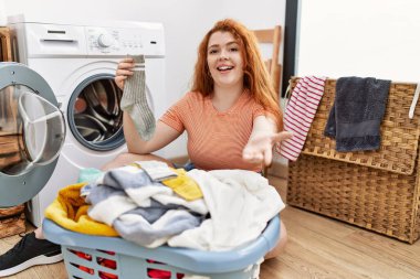 Kirli çamaşırları çamaşır makinesine koyan genç, kızıl saçlı bir kadın. Gülümseyen, neşeli bir el uzatan ve kabul gören bir kadın.. 