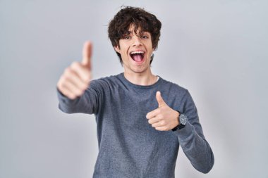 Young man standing over isolated background approving doing positive gesture with hand, thumbs up smiling and happy for success. winner gesture. 