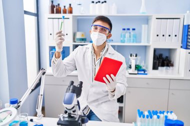 Latin kökenli genç bilim adamı laboratuarda dokunmatik ped kullanarak test tüpü tutuyor.