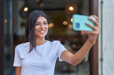 Genç ve güzel İspanyol kadın gülümsüyor. Kendine güveni tam. Sokakta akıllı telefonun yanında selfie çekiyor.