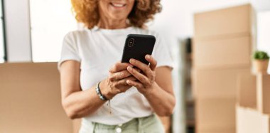 Orta yaşlı İspanyol kadın yeni evinde akıllı telefon kullanarak gülümsüyor.