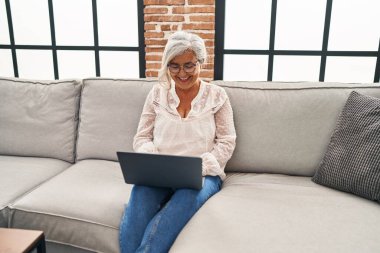 Orta yaşlı bir kadın evdeki koltukta dizüstü bilgisayar kullanıyor.