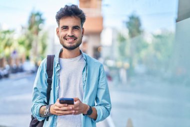 Latin asıllı genç bir öğrenci sokakta akıllı telefon kullanarak gülümsüyor.