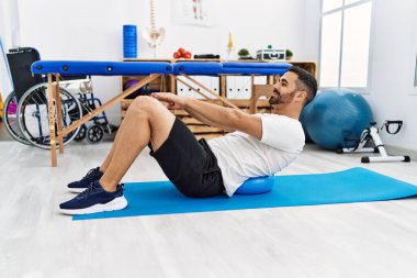 Young hispanic man patient smiling confident having back rehab session using fit ball at clinic