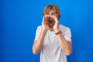 Orta yaşlı bir adam mavi arka planda duruyor ve elleri ağzında kızgın bir şekilde bağırıyor. 