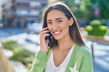 Genç, güzel, İspanyol bir kadın gülümsüyor ve caddede akıllı telefondan konuşuyor.