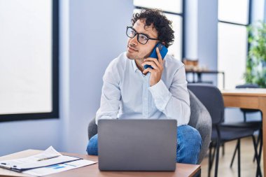 Young hispanic man business worker using laptop talking on smartphone at office