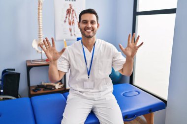 Sakallı, sakallı, acılı bir klinikte çalışan, on numaralı parmaklarıyla işaret eden, kendinden emin ve mutlu genç bir adam.. 