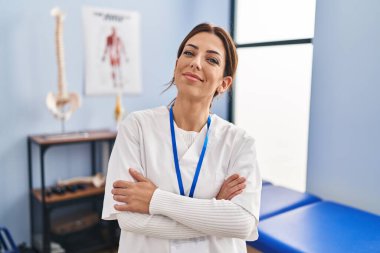 Acıyı dindirme kliniğinde çalışan genç esmer kadın kollarını kavuşturup kameraya bakarak gülümsüyor. pozitif kişi. 
