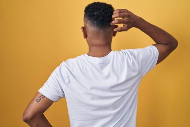 Young hispanic man standing over yellow background backwards thinking about doubt with hand on head 