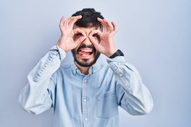 Mavi arka planda dikilen sakallı genç bir İspanyol dürbün gibi jest yapıyor, gözleri parmaklarından bakıyor. çılgın ifade. 