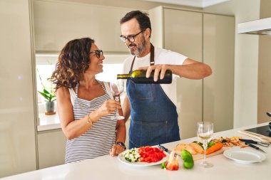 Middle age hispanic couple cooking and pouring wine on glass at kitchen