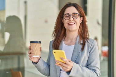 Sokak ortasında kahve içen akıllı telefon kullanan genç bir iş kadını.
