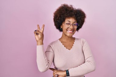 Genç Afro-Amerikan kadın pembe arka planda durup kameraya gülümseyen ve parmaklarıyla zafer işareti yapan. İki numara.. 