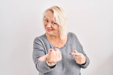 Beyaz arka planda dikilen orta yaşlı beyaz bir kadın buraya el hareketleriyle mutlu bir şekilde karşılamaya ve gülümsemeye davet ediyor. 