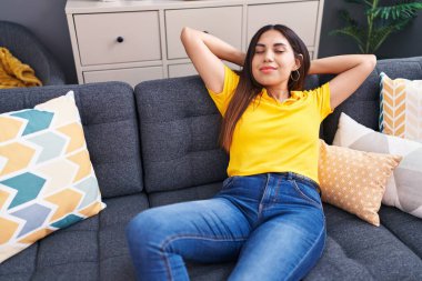 Young beautiful arab woman relaxed with hands on head sitting on sofa at home