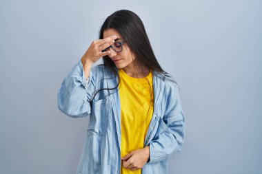 Mavi arka planda dikilen genç İspanyol kadın yorgun burnu ve gözleri ovuşturup yorgunluk ve baş ağrısı hissediyor. Stres ve hüsran kavramı. 