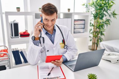 Young man doctor talking on smartphone using touchpad at clinic