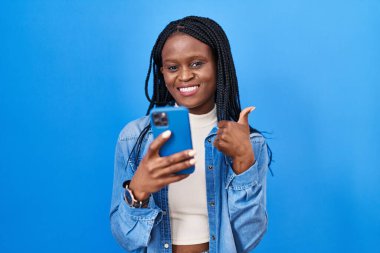 African woman with braids using smartphone typing message smiling happy and positive, thumb up doing excellent and approval sign 
