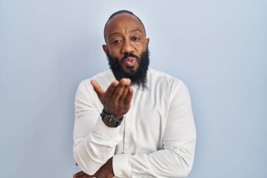 Afro-Amerikalı bir adam mavi arka planda durup kameraya bakarak havada bir öpücük savuruyor ve sevimli ve seksi oluyor. aşk ifadesi. 